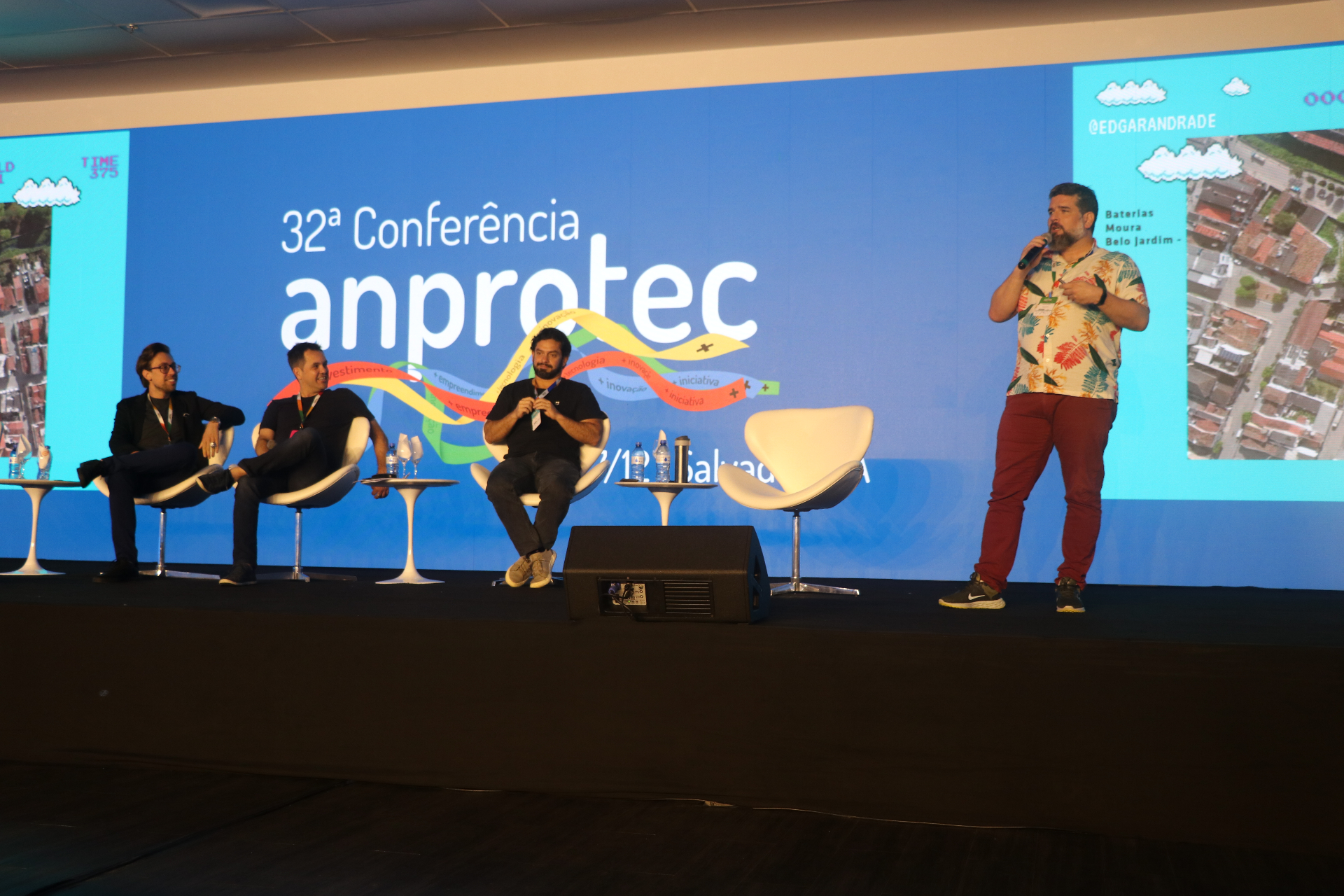 Edgar Andrade no palco da 32ª Conferência Anprotec, ao lado de painelistas em poltronas brancas, com telão azul ao fundo.
